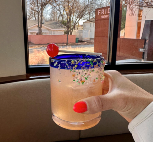 A hand holds a colorful cocktail with a cherry on top, set against a window view of a sunny outdoor scene.