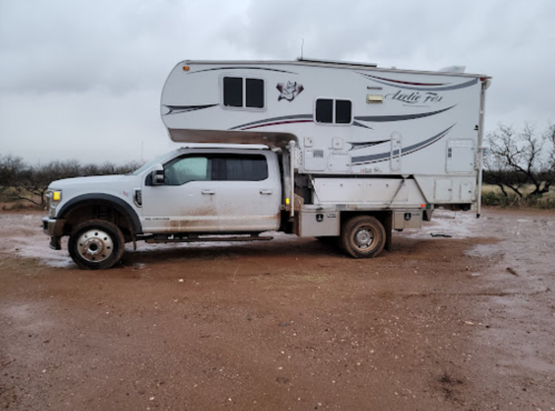 A white truck with a camper shell parked on a muddy, rural road under a cloudy sky.