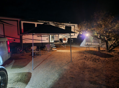 A campsite at night featuring a large RV, a tent, and a canopy with chairs under a tree.