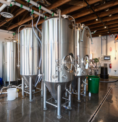 Three large stainless steel fermentation tanks in a brewery, with pipes and equipment visible in a well-lit space.