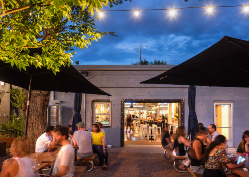 Outdoor dining area with string lights, people enjoying meals, and a cozy atmosphere under large umbrellas.