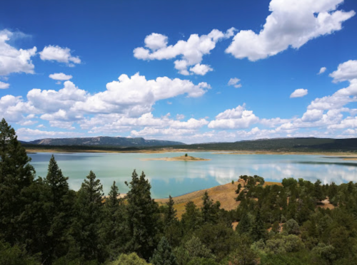 A serene lake surrounded by trees under a blue sky with fluffy clouds, reflecting the landscape in calm waters.