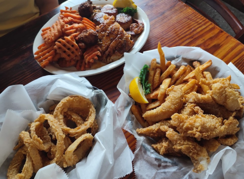 A table with three baskets of food: fried fish, onion rings, and fries, alongside a plate of grilled chicken and sweet potato fries.