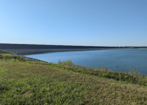 A calm body of water beside a grassy shore, with a large, dark embankment in the background under a clear blue sky.