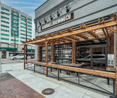 Exterior of 10 Barrel Brewing Co. featuring a wooden patio and large windows, with a cityscape in the background.