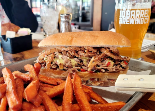 A hearty sandwich filled with meat and toppings, served with a side of sweet potato fries and a glass of beer.