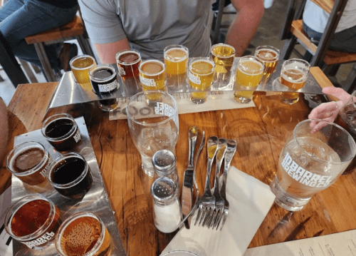 A wooden table with a flight of beer samples and glasses of water, alongside utensils and a menu.
