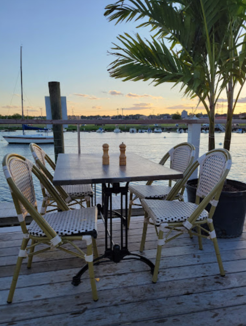 A waterfront dining area with a table and chairs, overlooking boats and a sunset. Palm trees in the background.