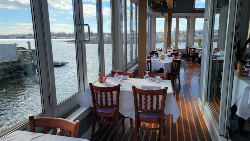 A waterfront restaurant with tables set for dining, large windows offering a view of the water and sky.