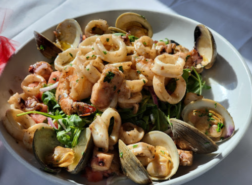 A bowl of seafood pasta featuring shrimp, calamari, and clams on a bed of greens, garnished with herbs.