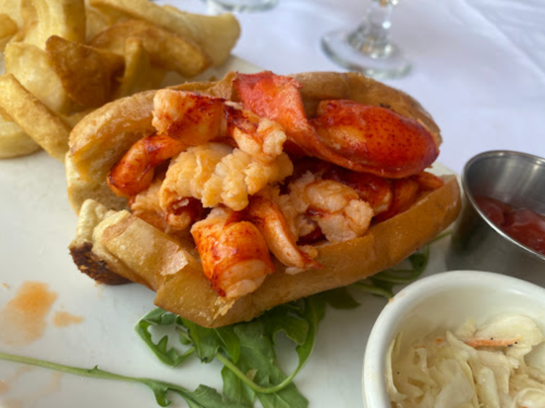 A lobster roll filled with chunks of lobster meat, served with fries and sides of coleslaw and cocktail sauce.