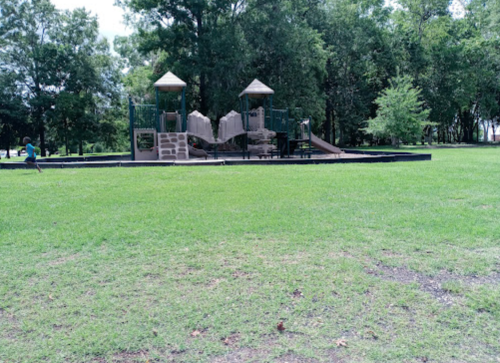 A playground with slides and climbing structures in a grassy park surrounded by trees.