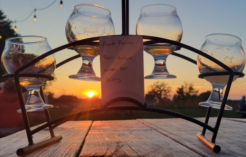 Four glasses on a stand with a note, set against a sunset backdrop. Warm colors and a blurred landscape in the distance.
