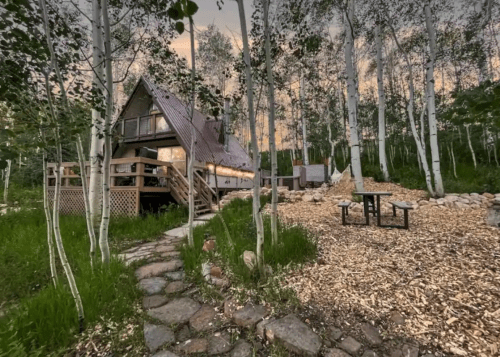 A cozy A-frame cabin surrounded by aspen trees, with a stone path and picnic area in a serene outdoor setting.