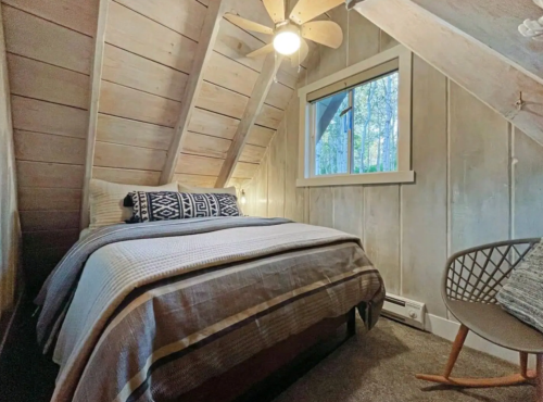 Cozy attic bedroom with a double bed, wooden walls, and a window overlooking trees. A fan and chair complete the space.