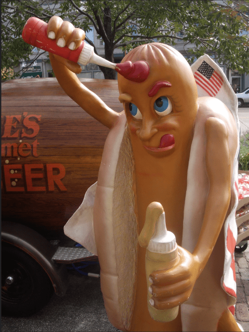 A cartoonish hot dog character with a bottle, playfully squeezing ketchup, next to a wooden beer barrel.