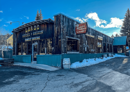 A rustic wooden building with signs for "Lardo Grill & Saloon" and "Family Dining," surrounded by snow and trees.
