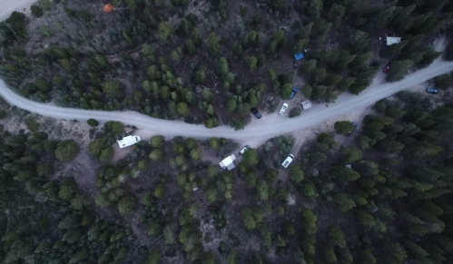 Aerial view of a winding dirt road surrounded by dense trees, with several parked vehicles visible along the route.