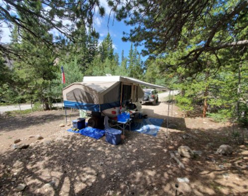 A campsite with a pop-up camper surrounded by trees, a picnic area, and a gravel road in the background.