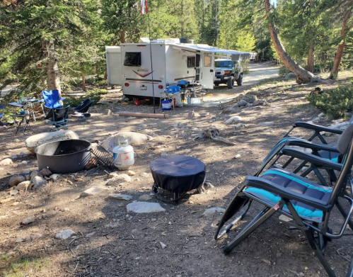 A campsite with a trailer, chairs, and a fire pit surrounded by trees and rocky ground.
