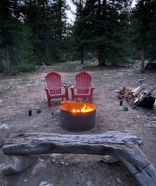 Two red chairs beside a fire pit with flames, surrounded by trees and firewood in a serene outdoor setting.