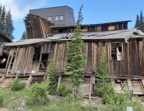 A dilapidated wooden building leaning at an angle, surrounded by trees and a blue sky.