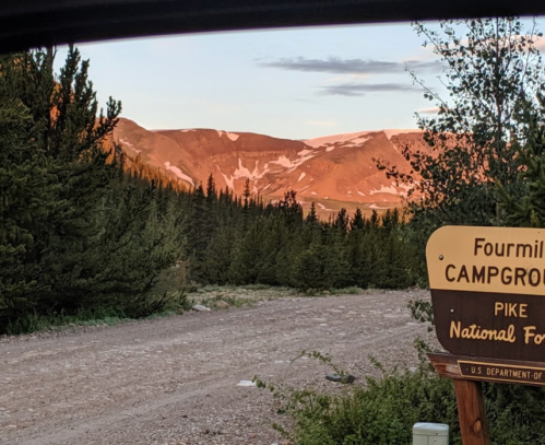 A scenic view of mountains at sunset, framed by trees, with a sign for Fourmile Campground in the foreground.