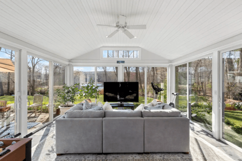 Bright sunroom with a gray sofa, large windows, a TV, and views of a garden outside.