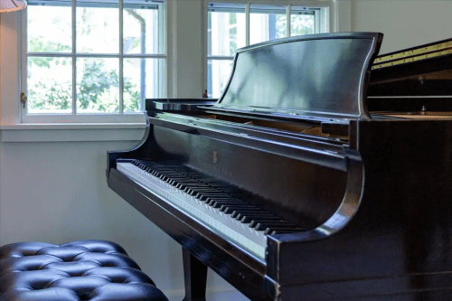 A grand piano beside a window, with sunlight illuminating its polished surface and a black tufted bench nearby.