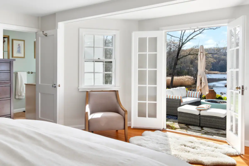 Bright bedroom with a chair, open doors leading to a patio by a river, and natural light streaming in.