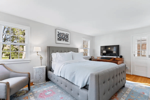 Bright bedroom featuring a gray tufted bed, a TV, a cozy armchair, and large windows with a view of greenery.