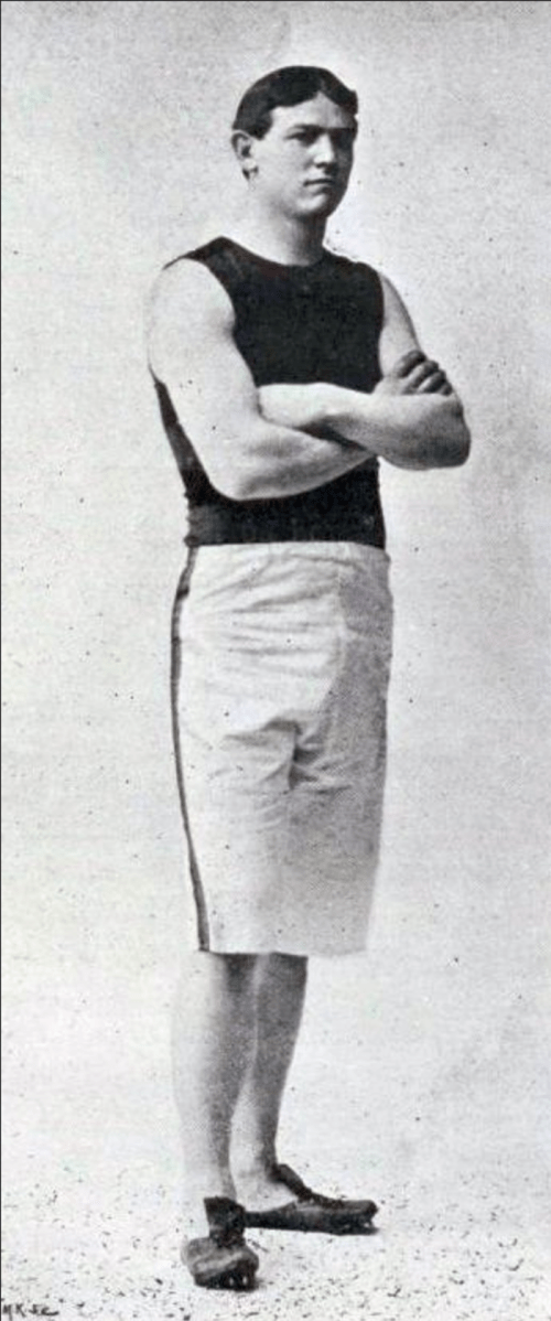 A vintage black and white photo of a man in athletic wear, standing with arms crossed, looking confidently at the camera.