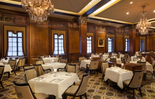 Elegant restaurant interior with wooden paneling, chandeliers, and neatly set tables. Warm, inviting atmosphere.