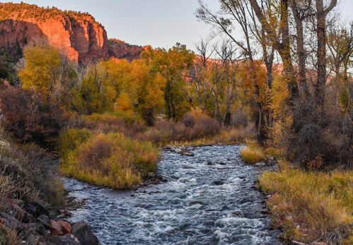 A serene river flows through a colorful autumn landscape with trees and red rock formations in the background.
