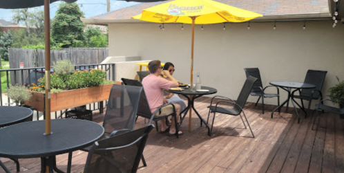 A couple sits at a table on a wooden patio, enjoying a meal under a yellow umbrella.