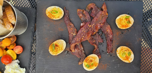 A slate platter featuring strips of beef jerky, deviled eggs, and a side of colorful cherry tomatoes and bread.