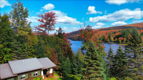 A cozy cabin surrounded by colorful autumn trees near a serene lake under a bright blue sky with fluffy clouds.