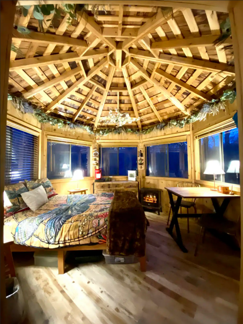 Cozy wooden cabin interior with a circular ceiling, large windows, a bed, and a small dining area. Warm lighting creates a welcoming atmosphere.