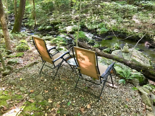 Two chairs by a serene stream in a wooded area, with a small statue nearby and lush greenery surrounding the scene.