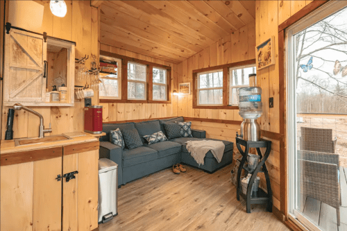 Cozy wooden interior of a tiny home featuring a sofa, kitchenette, and large windows with a view of nature outside.