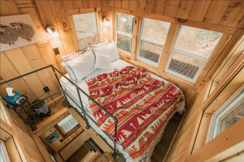 Cozy wooden cabin interior with a bed, colorful blanket, and large windows, featuring a staircase leading down.