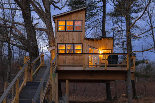 A cozy wooden treehouse with glowing windows, elevated on a deck surrounded by trees at dusk.