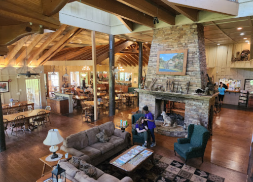 A spacious lodge interior with a stone fireplace, wooden beams, and dining area, featuring cozy seating and rustic decor.