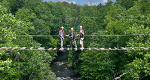 Three people in helmets stand on a suspension bridge surrounded by lush green trees and a river below.