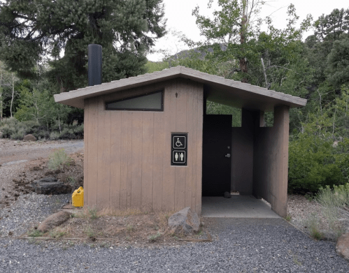 A small, accessible restroom building in a natural setting, surrounded by trees and gravel pathways.
