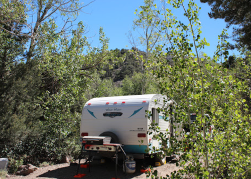 A small white camper surrounded by trees and greenery, set against a backdrop of mountains under a clear blue sky.