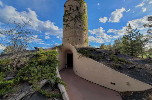 A stone tower covered in vines, surrounded by greenery and a blue sky with clouds, leading to a curved entrance.