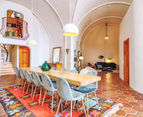 A spacious dining area with a long wooden table, stylish chairs, and a cozy living space in a modern, arched room.