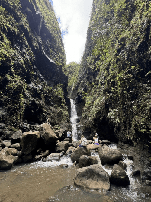 A serene waterfall cascades between steep, rocky cliffs, with people sitting on large stones by the water's edge.