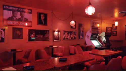 A retro bar interior with pink lighting, featuring vintage photos, arcade games, and cozy seating.